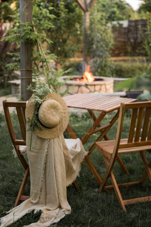 Foto de Muebles de madera en el jardín contra el telón de fondo de un fuego en el patio. Entre las ramas del arbusto se encuentra una mesa de madera y dos sillas de madera. Uno de ellos lleva un sombrero de paja y una manta a cuadros. Chimenea en la distancia en forma de una belleza - Imagen libre de derechos