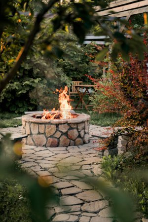 Photo for Patio and fire in the fire pit in the evening. Beautiful fire flames surrounded by beautiful trees. In the background is a table and two chairs. Summer collection - Royalty Free Image