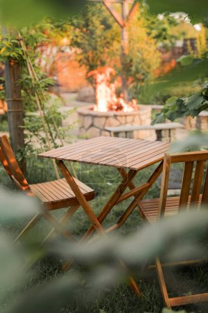 Foto de Muebles de madera en el jardín contra el telón de fondo de un fuego en el patio. Entre las ramas del arbusto se encuentra una mesa de madera y sillas. Vertical - Imagen libre de derechos