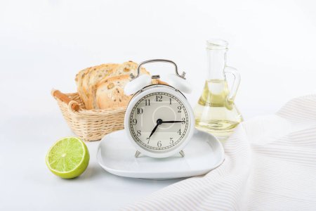 Foto de Reloj despertador blanco sobre un plato blanco sobre el fondo de una botella de aceite, pan y lima. Es la hora del desayuno en el despertador. Concepto de dieta. Menú desayuno - Imagen libre de derechos