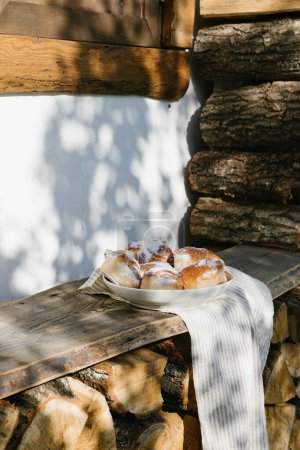 The pies in the plate stand on a wooden bench outside the village house. Next to the house is a large beautiful woodpile. There are nice shadows from the trees on the white wall. Rural scene of summer