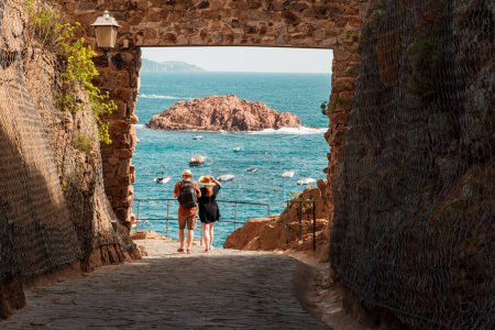 Photo for Tourists at the gate to the Villa Vella fortress in Tossa de Mar, Catalonia, Spain. The Bay of Badia di Tossa and the island L'Illa in the background - Royalty Free Image