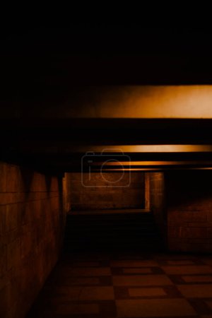 Un oscuro túnel sombreado iluminado por unas pequeñas lámparas que esparcen luz amarilla sobre las baldosas. Un cruce peatonal subterráneo