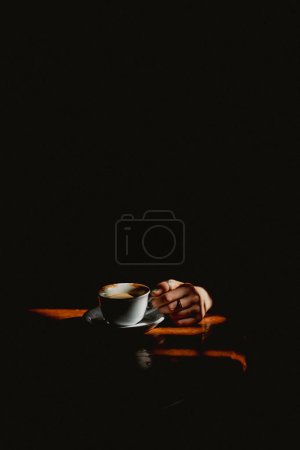 Eine Kaffeetasse, die auf einem Holztisch steht und von der warmen Sommersonne erhellt wird, und ein Mädchen, das im Schatten sitzt und sie hält. Ein Konzept von Mysterien, Geheimnissen, Identität und Mystikern.