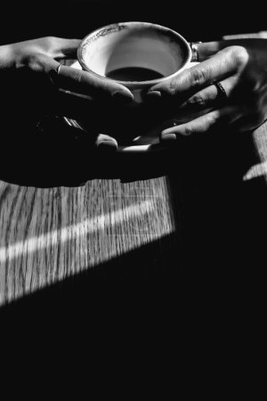 Foto de Una chica sosteniendo una taza de café de pie sobre una mesa de estilo vintage. Imagen misteriosa con café negro y sombras oscuras con un toque negro. - Imagen libre de derechos