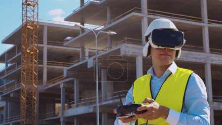 Téléchargez les photos : Opérateur de drone professionnel en réalité virtuelle casque debout devant le chantier de construction. Builder tenant la télécommande. Bâtiment de bureaux et fond de grue. Affaires, immobilier et - en image libre de droit