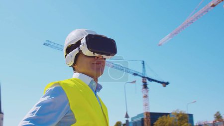 Téléchargez les photos : Constructeur professionnel en casque VR debout devant le chantier de construction et en utilisant des technologies de réalité virtuelle et augmentée. Bâtiment de bureaux et fond de grue. Immobilier et concepts d'investissement - en image libre de droit