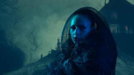 Schöne Hexe, die die Hexerei über dem rauchigen Hintergrund in der Nacht macht. Gruseliges Haus auf dem Hügel. Imagekonzept für Halloween.