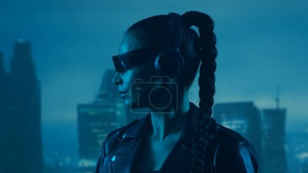 Téléchargez les photos : Portrait de cyberpunk girl avec un smartphone dans des lunettes et des écouteurs. Belle jeune femme sur le fond des grattoirs de la ville. Concept futuriste. - en image libre de droit