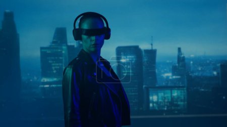 Photo for Portrait of cyberpunk girl with a smartphone in a glasses and headphones. Beautiful young woman on the background of city scyscrapers. Futuristic concept. - Royalty Free Image