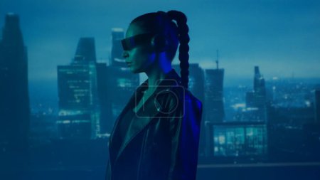 Téléchargez les photos : Portrait de cyberpunk girl avec un smartphone dans des lunettes et des écouteurs. Belle jeune femme sur le fond des grattoirs de la ville. Concept futuriste. - en image libre de droit
