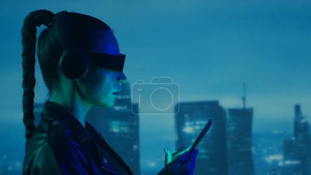 Foto de Retrato de chica cyberpunk con un smartphone en gafas y auriculares. Hermosa joven en el fondo de rascacielos de la ciudad. Concepto futurista. - Imagen libre de derechos