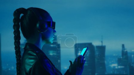 Foto de Retrato de chica cyberpunk con un smartphone en gafas y auriculares. Hermosa joven en el fondo de rascacielos de la ciudad. Concepto futurista. - Imagen libre de derechos