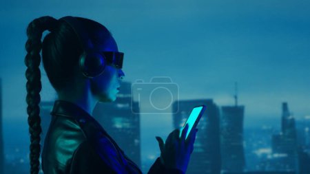 Photo for Portrait of cyberpunk girl with a smartphone in a glasses and headphones. Beautiful young woman on the background of city scyscrapers. Futuristic concept. - Royalty Free Image