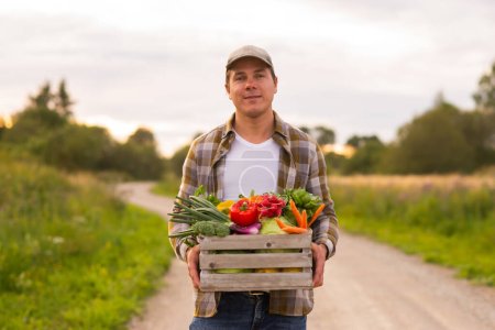 Téléchargez les photos : Farmer with a vegetable box in front of a sunset agricultural landscape. Man in a countryside field. The concept of country life, food production, farming and country lifestyle. - en image libre de droit