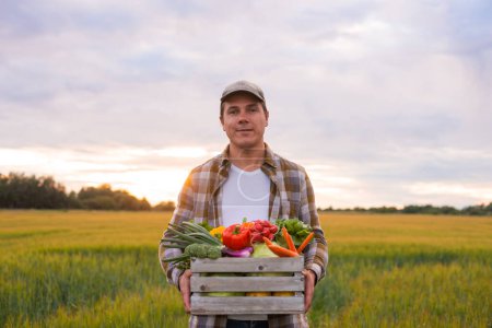 Foto de Farmer with a vegetable box in front of a sunset agricultural landscape. Man in a countryside field. The concept of country life, food production, farming and country lifestyle. - Imagen libre de derechos