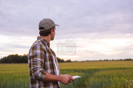 Téléchargez les photos : Farmer with a tablet computer in front of a sunset agricultural landscape. Man in a countryside field. The concept of country life, food production, farming and technology. - en image libre de droit