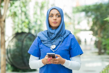 Téléchargez les photos : Close-up portrait of young and attractive muslim woman nurse in hijab. Middle Eastern female doctor outdoor on the street. City background. The concept of medicine and health care. - en image libre de droit