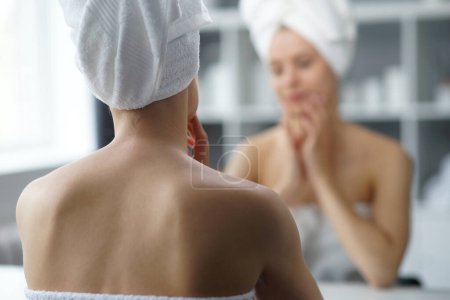 Téléchargez les photos : Young woman sits in the bathroom in front of the makeup mirror and does cosmetic procedures. Beautiful girl in white towel. The concept of skin care, health, rejuvenation and spa treatment. - en image libre de droit