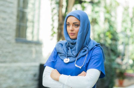 Foto de Close-up portrait of young and attractive muslim woman nurse in hijab. Middle Eastern female doctor outdoor on the street. City background. The concept of medicine and health care. - Imagen libre de derechos