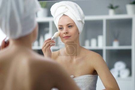 Foto de Young woman sits in the bathroom in front of the makeup mirror and does cosmetic procedures. Beautiful girl in white towel. The concept of skin care, health, rejuvenation and spa treatment. - Imagen libre de derechos