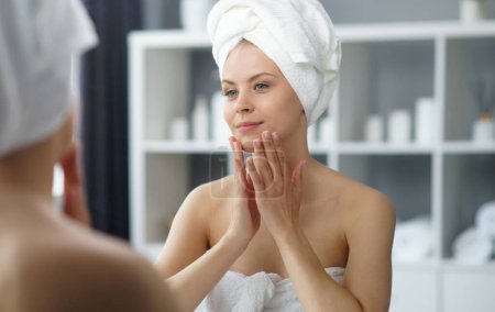 Téléchargez les photos : Young woman sits in the bathroom in front of the makeup mirror and does cosmetic procedures. Beautiful girl in white towel. The concept of skin care, health, rejuvenation and spa treatment. - en image libre de droit