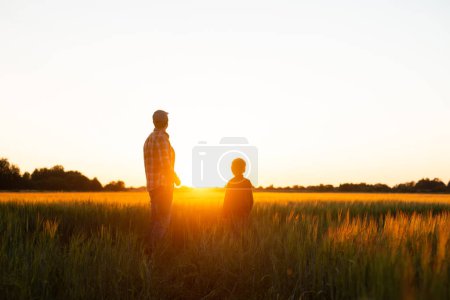 Téléchargez les photos : Farmer and his son in front of a sunset agricultural landscape. Man and a boy in a countryside field. The concept of fatherhood, country life, farming and country lifestyle. - en image libre de droit