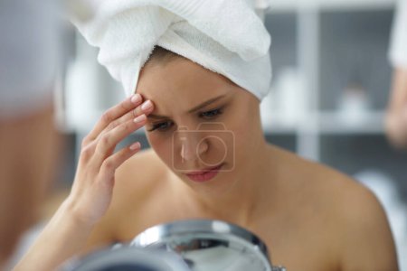 Foto de Young woman sits in the bathroom in front of the makeup mirror and does cosmetic procedures. Beautiful girl in white towel. The concept of skin care, health, rejuvenation and spa treatment. - Imagen libre de derechos