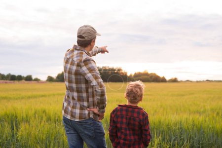 Foto de Farmer and his son in front of a sunset agricultural landscape. Man and a boy in a countryside field. The concept of fatherhood, country life, farming and country lifestyle. - Imagen libre de derechos
