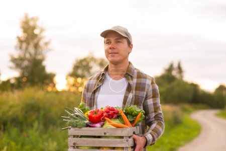 Photo for Farmer with a vegetable box in front of a sunset agricultural landscape. Man in a countryside field. The concept of country life, food production, farming and country lifestyle. - Royalty Free Image
