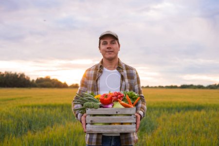 Photo for Farmer with a vegetable box in front of a sunset agricultural landscape. Man in a countryside field. The concept of country life, food production, farming and country lifestyle. - Royalty Free Image