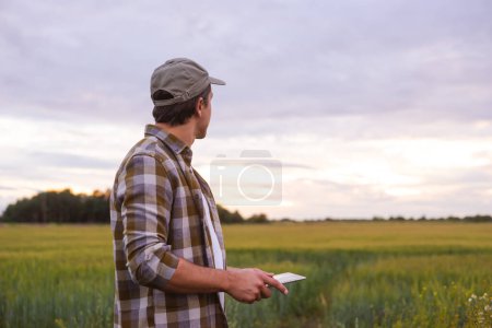 Téléchargez les photos : Farmer with a tablet computer in front of a sunset agricultural landscape. Man in a countryside field. The concept of country life, food production, farming and technology. - en image libre de droit