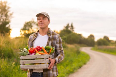 Téléchargez les photos : Farmer with a vegetable box in front of a sunset agricultural landscape. Man in a countryside field. The concept of country life, food production, farming and country lifestyle. - en image libre de droit