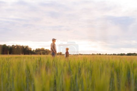 Photo for Farmer and his son in front of a sunset agricultural landscape. Man and a boy in a countryside field. The concept of fatherhood, country life, farming and country lifestyle. - Royalty Free Image