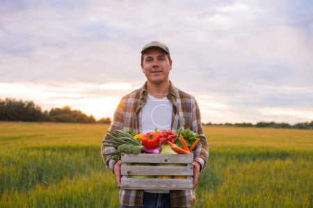 Foto de Farmer with a vegetable box in front of a sunset agricultural landscape. Man in a countryside field. The concept of country life, food production, farming and country lifestyle. - Imagen libre de derechos