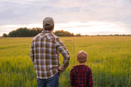 Foto de Farmer and his son in front of a sunset agricultural landscape. Man and a boy in a countryside field. The concept of fatherhood, country life, farming and country lifestyle. - Imagen libre de derechos