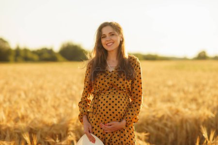 Photo for Pregnant woman in the rays of the sunset. Beautiful young girl is walking in the field expecting the birth of a child. The concept of motherhood, pregnancy and happiness. - Royalty Free Image