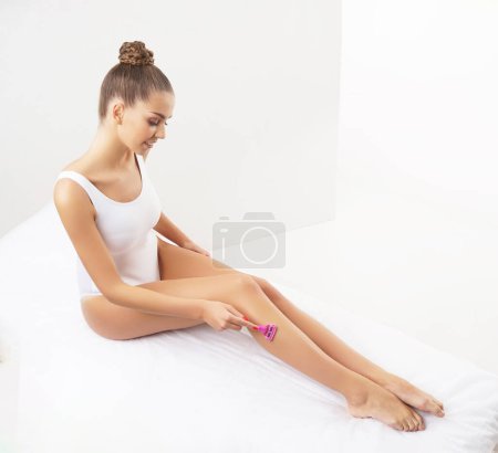 Photo for Young, fit and beautiful brunette woman in white swimsuit doing epilation over white background. Healthcare, body care, depilation and spa concept. - Royalty Free Image
