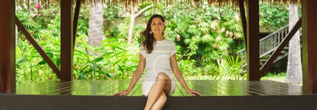 Photo for Young and beautiful woman in white dress. Woman posing in exotic Thai bungalow. Resting and traveling concept. Jungle background. - Royalty Free Image