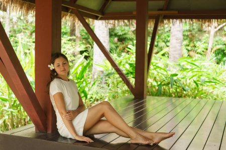 Téléchargez les photos : Jeune et belle femme en robe blanche. Femme posant dans un bungalow thaïlandais exotique. Concept de repos et de voyage. Fond de jungle. - en image libre de droit