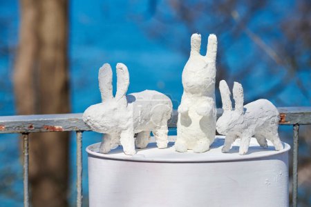 Foto de Three white rabbit statues made of plaster on barrel at outdoor art exhibition, artificial white hares. White handmade bunnies, artificial rabbits outdoor, Easter urban decor concept - Imagen libre de derechos