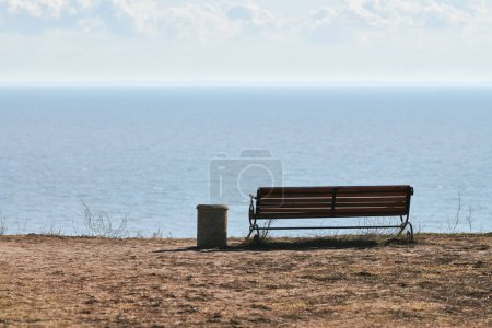 Foto de Empty bench with trashcan on cliff before sea background, peaceful and quiet place for thinking alone, loneliness and loss of loved one concept. Pacifying view of marine horizon of Azov sea copy space - Imagen libre de derechos