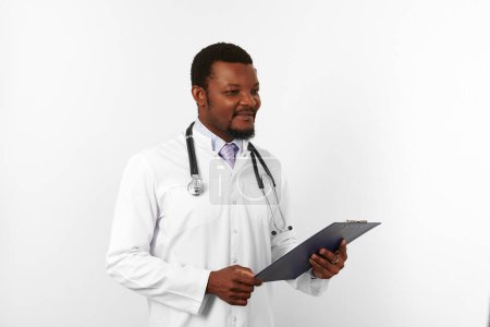 Smiling black bearded doctor man in white robe with stethoscope holds medical chart on clipboard, isolated on white background. Happy adult black african american physician therapist portrait