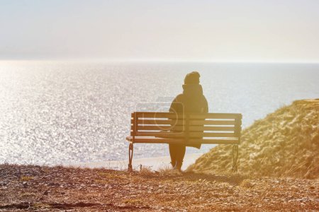 Foto de Single girl in a black jacket and hat sitting on bench at cliff at front of sea, peaceful and quiet place for thinking alone, loneliness and loss of loved one concept. Pacifying view of marine horizon - Imagen libre de derechos