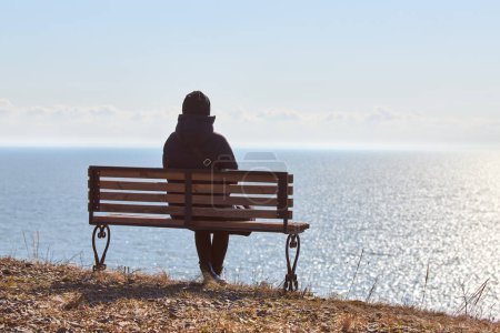 Foto de Single girl in a black jacket and hat sitting on bench at cliff at front of sea, peaceful and quiet place for thinking alone, loneliness and loss of loved one concept. Pacifying view of marine horizon - Imagen libre de derechos