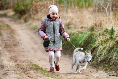 Photo for Girl walking on leash Siberian Husky puppy on country road at forest background, happy friendship of dog and cute little kid. Young girl running Husky dog breed puppy on autumn outdoor road - Royalty Free Image