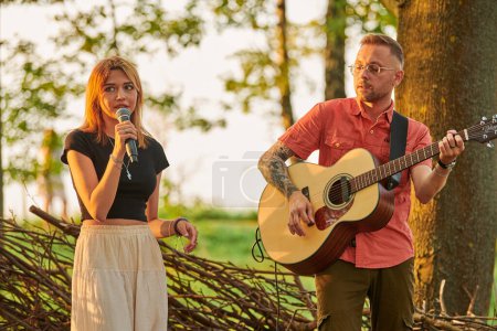 Photo for Svetlogorsk, Russia - 2022.08.14 - Young girl singer holds microphone in hand with man guitarist plays pop rock music at outdoor art festival, green forest background. Summer rock band performance - Royalty Free Image