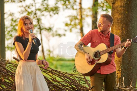 Photo for Svetlogorsk, Russia - 2022.08.14 - Young girl singer holds microphone in hand with man guitarist plays pop rock music at outdoor art festival, green forest background. Summer rock band performance - Royalty Free Image