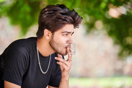 Photo for Young indian man smoker portrait in black t shirt and silver neck chain in public park, hindu male smoking close up portrait. Handsome indian man portrait with thick hair in city green park - Royalty Free Image