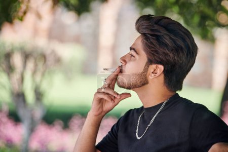 Photo for Young indian man smoker portrait in black t shirt and silver neck chain sitting on bench in public park, hindu male smoking close up portrait. Handsome indian man portrait with thick hair in city park - Royalty Free Image